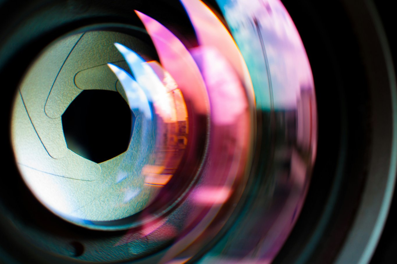 Extreme close-up of a camera lens aperture blades forming a hexagonal opening, ringed with pink and cyan flare