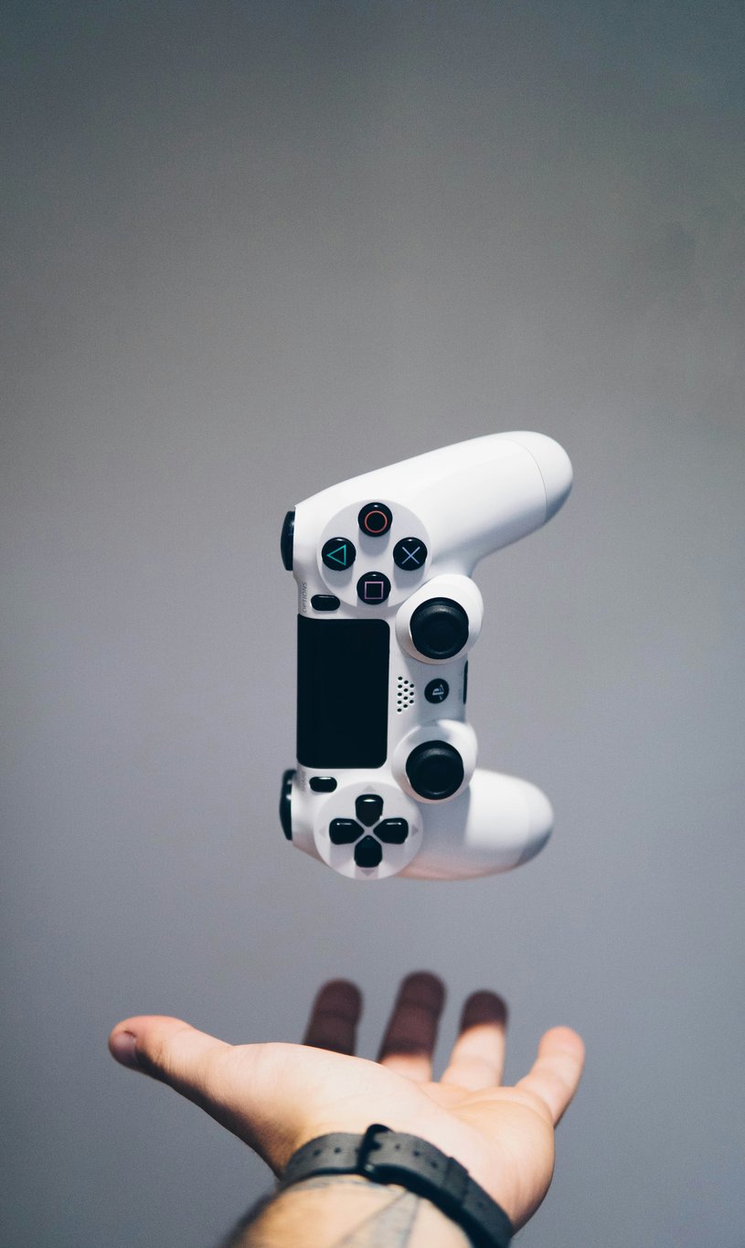 An open palm held beneath a white games controller suspended in mid-air against a grey wall