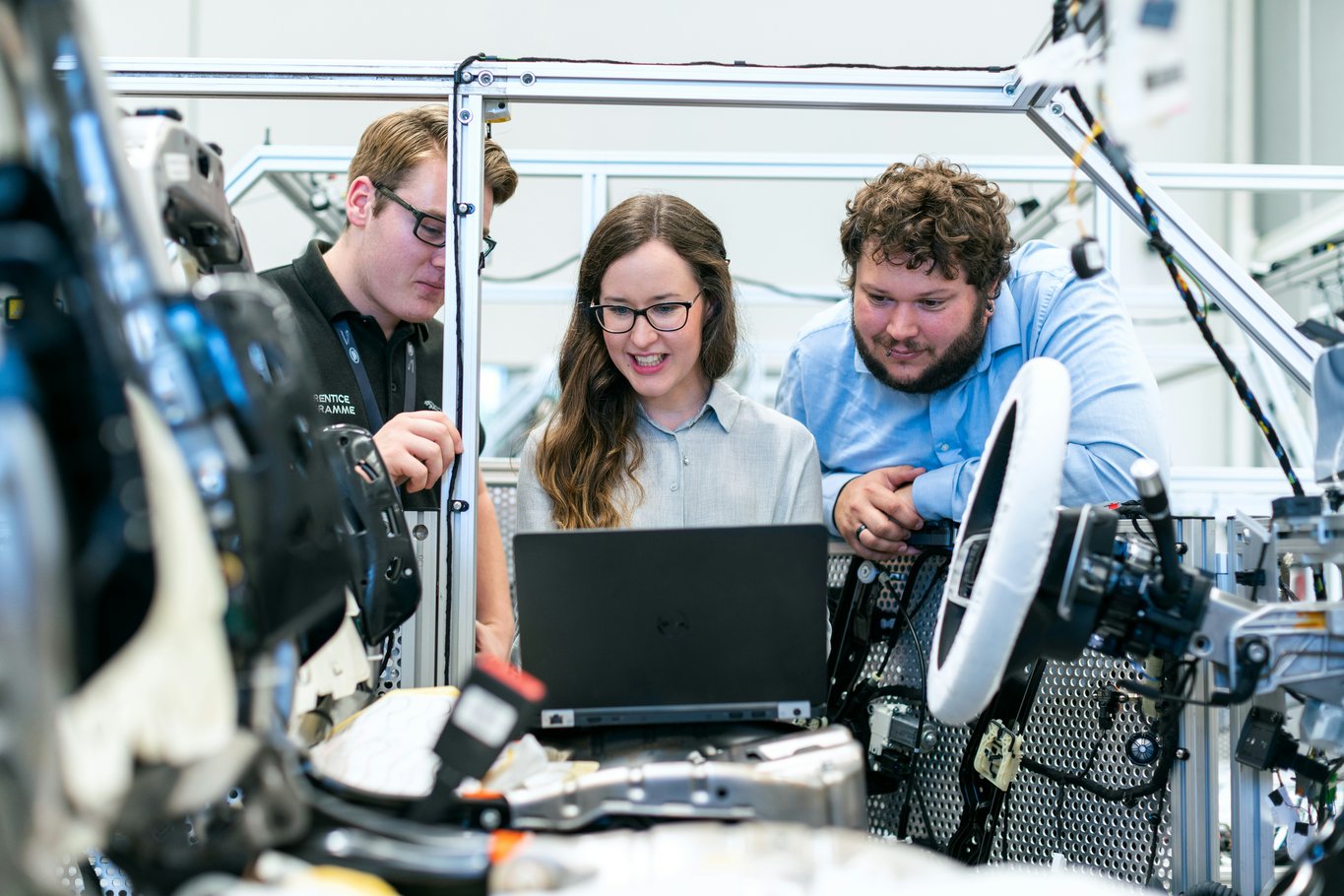 Three engineers in a bright laboratory leaning over a laptop beside a partially disassembled vehicle test rig