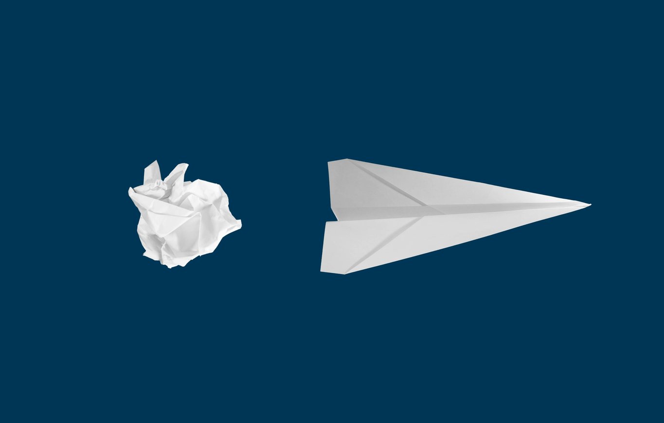A crumpled white paper ball beside a neatly folded paper aeroplane on a deep navy background