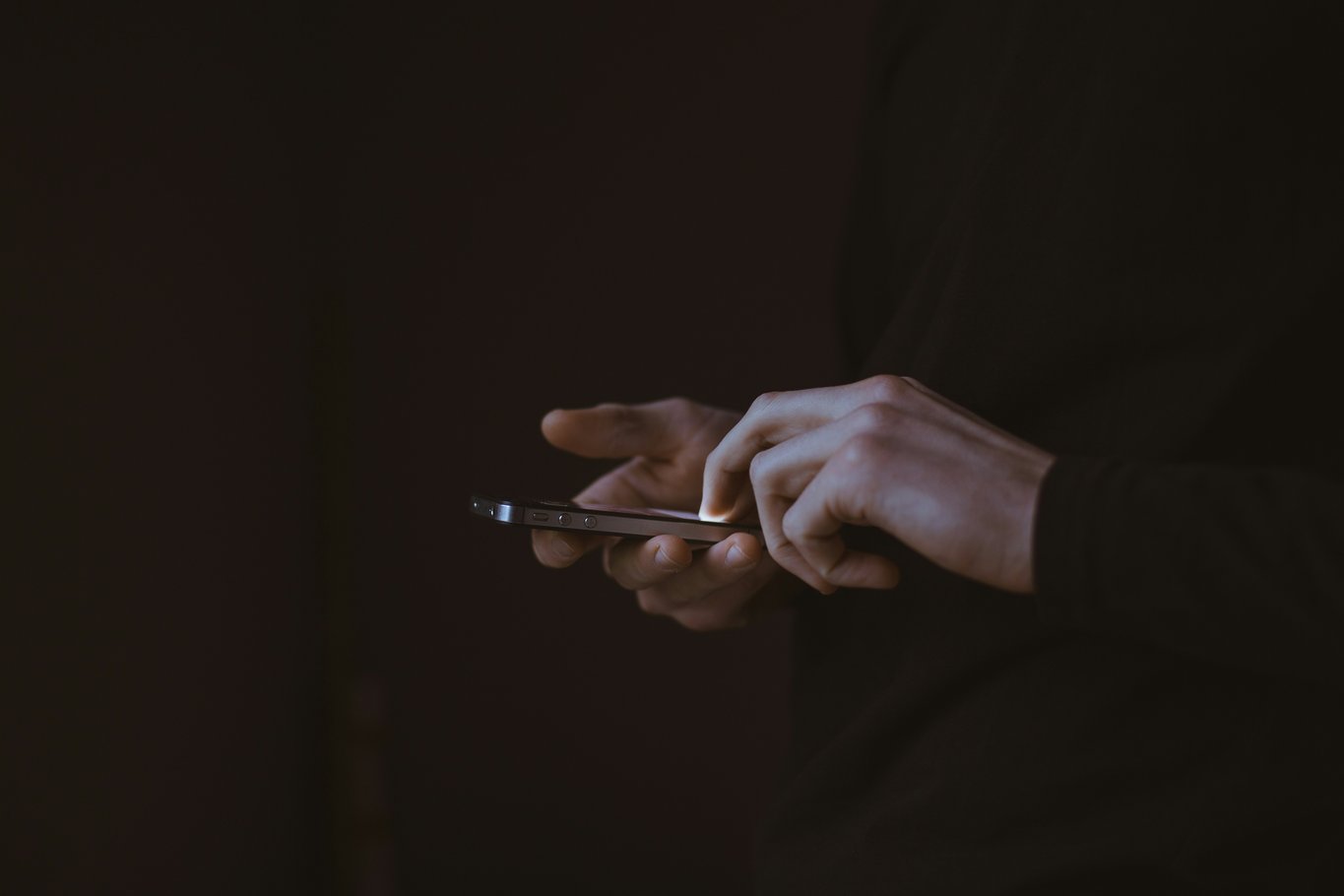Two hands holding and tapping a dark smartphone in near-darkness, lit only by the screen