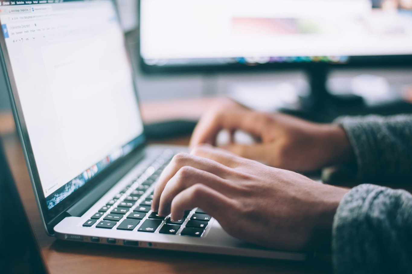 Close-up of hands typing on a silver laptop keyboard at a wooden desk with a blurred monitor behind
