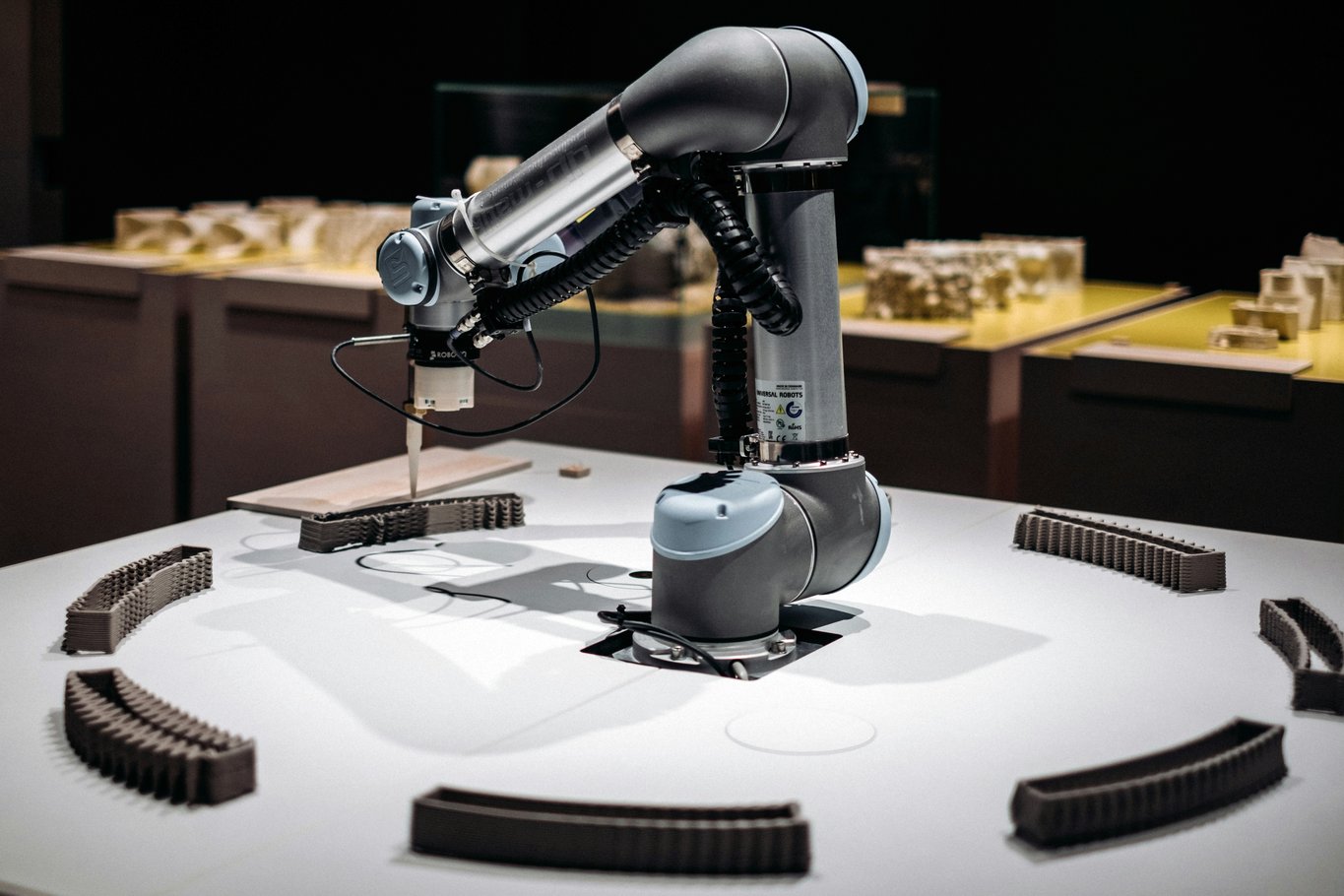 An industrial robotic arm on a white table extruding material beside curved ridged printed components