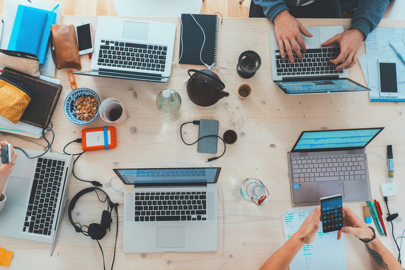 Overhead view of a pale wooden desk with four open laptops, several pairs of hands typing, notebooks and cups
