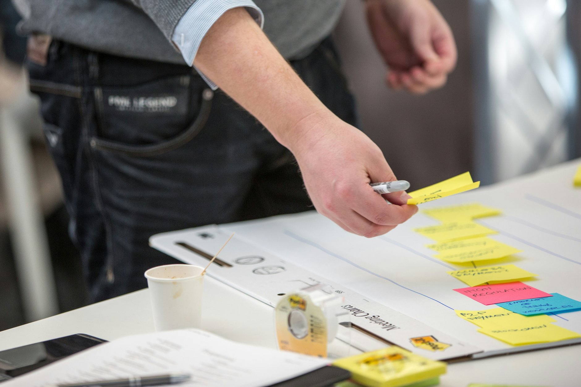 A hand adding a yellow sticky note to a large sheet already covered in rows of handwritten coloured notes
